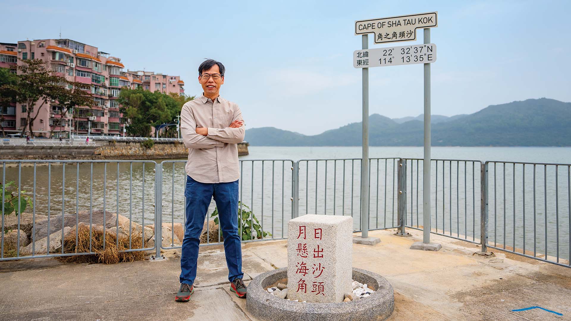 李以强站在沙头角之角的旧式路牌前，身后围绕着沙头角海湾的天然海岸线。